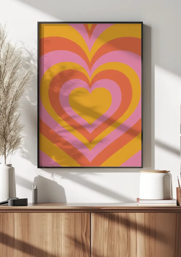 Een ingelijst Harten Patroon Schilderij van CollageDepot met een kleurrijk, concentrisch hartontwerp met roze, oranje en gele tinten wordt aan een witte muur boven een houten kast gehangen. Links van de wanddecoratie staat een vaas met gedroogd gras en rechts diverse potjes en een kaars.,Zwart