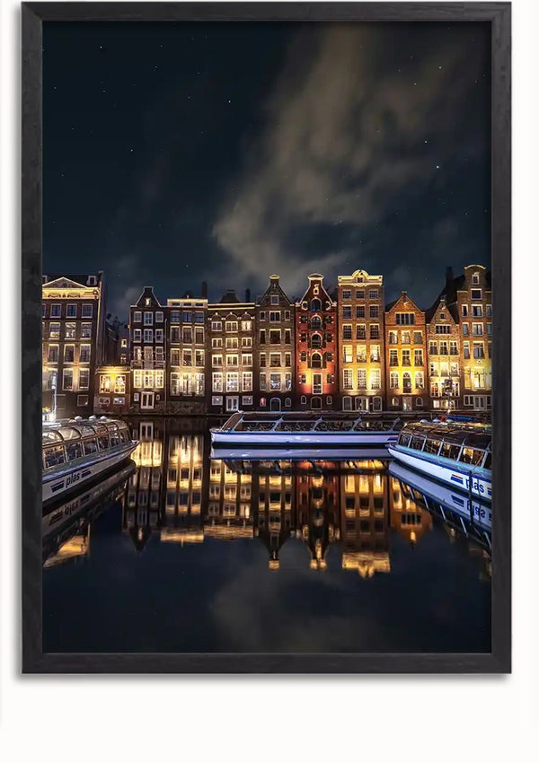 Een nachtelijk tafereel van een kanaal met rivierboten aangemeerd voor een rij verlichte, traditionele Amsterdamse grachtenpanden, weerspiegeld in het water. De lucht is donker met enkele verspreide wolken, waardoor een betoverend Amsterdamse Grachtenpanden Schilderij van CollageDepot ontstaat.,Lichtbruin