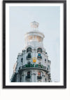 Een ingelijste foto, die doet denken aan een schilderij, toont een wit gebouw met meerdere verdiepingen, een ronde toren en verschillende balkons. Het gebouw beschikt over meerdere ramen en een sierlijk architectonisch ontwerp. Op de benedenverdieping zijn het logo en de naam van een bekend horlogemerk weergegeven. Dit is het **Rolex winkel Madrid schilderij** van **CollageDepot**.