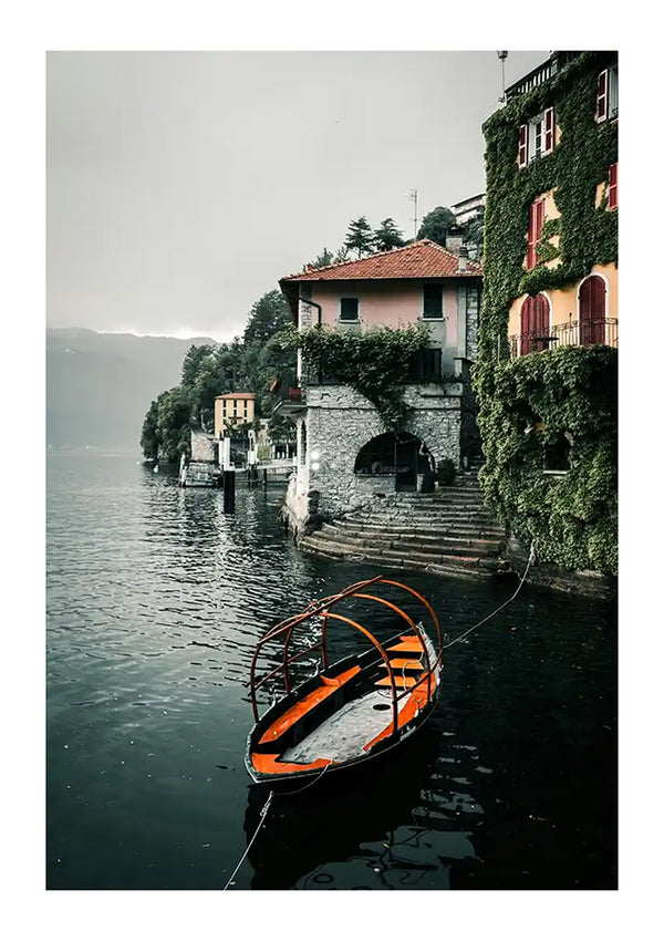 Een oranje en zwarte roeiboot ligt afgemeerd aan de rand van een waterkant. Achter de boot staan gebouwen met met klimop bedekte muren, trappen die naar het water leiden en een mistige achtergrond met bomen; het is als een levend kunstwerk, perfect voor wanddecoratie. Dit pittoreske tafereel is prachtig vastgelegd in het Place at Comolake-schilderij van CollageDepot.