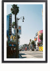 Een ingelijste foto, perfect voor wanddecoratie, toont een druk straatbeeld in Hollywood, Californië. Palmbomen omzomen de straat, met diverse winkels en reclameborden zichtbaar. Op een verticaal bord staat 'Hollywood' op de zijkant van een gebouw. Auto's en voetgangers dragen bij aan de levendige sfeer. Dit is het Hollywood Boulevard-schilderij van CollageDepot.,Zwart-Met,Lichtbruin-Met,showOne,Met