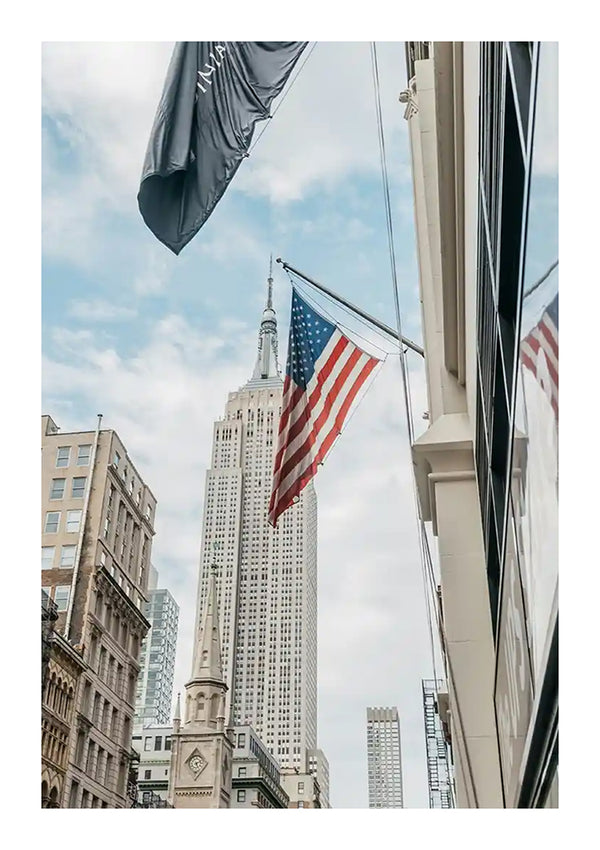 Op een foto genomen vanaf straatniveau, naar boven kijkend, is een Amerikaanse vlag te zien die aan een gebouw hangt met het Empire State Building en andere wolkenkrabbers op de achtergrond. De gedeeltelijk bewolkte hemel omlijst extra stadsgebouwen langs de straat, wat een gevoel van iconische New York City-charme oproept: een perfect New York City-schilderij van CollageDepot.-