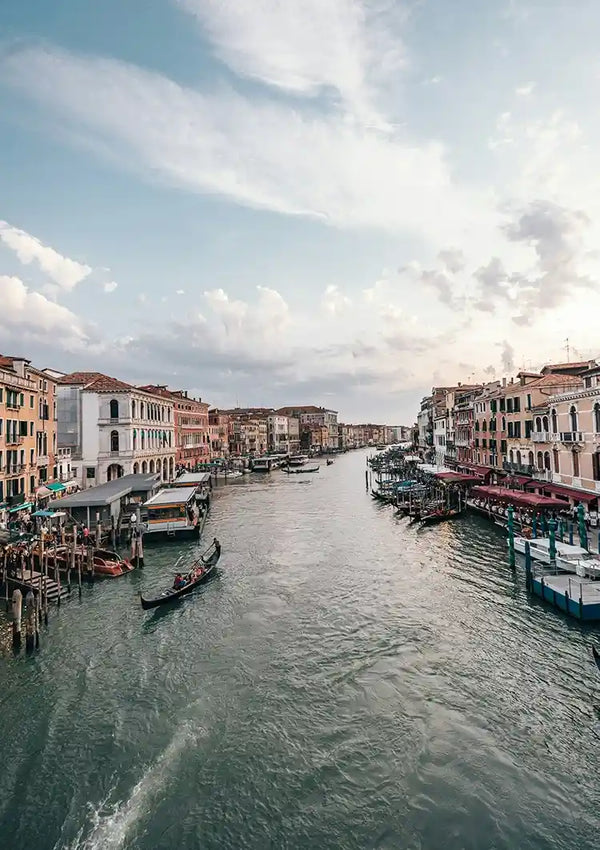 Een zicht op een kanaal in Venetië, Italië, met gondels en boten op het water. De gracht wordt geflankeerd door historische gebouwen, waarbij verschillende kleuren en architectonische details zichtbaar zijn. De lucht erboven is gedeeltelijk bewolkt. Dit Canal Grande Schilderij van CollageDepot zorgt voor prachtige wanddecoratie met zijn magnetische ophangsysteem.-