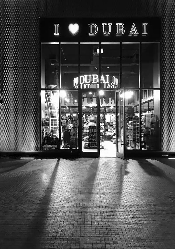 Een zwart-witfoto toont de hoofdingang van een winkel met "I ♥ Dubai" verlicht boven de deuren. In de helder verlichte winkel zijn I love Dubai-schilderijitems van CollageDepot zichtbaar. De gevel van de winkel heeft een muur met patronen en de grond buiten is geplaveid met bakstenen.