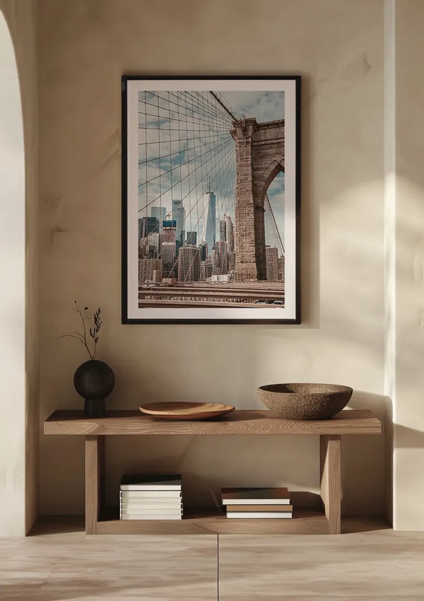 Het minimalistische interieur bestaat uit een lichthouten consoletafel tegen een beige muur. Een Uitzicht Skyline New York Schilderij van CollageDepot hangt erboven door middel van een magnetisch ophangsysteem. Op de tafel staan een vaas met een takje, een ondiepe kom, een diepe kom en gestapelde boeken. Zonlicht werpt schaduwen op het toneel.,Zwart