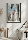 Een ingelijste foto van een stedelijke scène met een stadsgebouw met het Empire State Building op de achtergrond, met een New York City Schilderij van CollageDepot. Hieronder staat een lichtgekleurde houten kruk met een boek en een plantje, en een stoel met een gedrapeerde witte deken.,Zwart