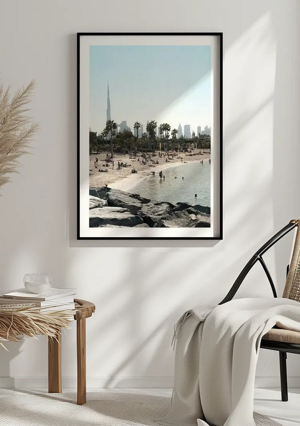 Een ingelijst CollageDepot Skyline Dubai Schilderij van een strandtafereel wordt aan een gebroken witte muur gehangen, met rechts een knusse stoel gedrapeerd in een lichte deken. Links van de stoel staat een kleine, ronde houten tafel met een paar spullen erop, en gedroogde planten dragen bij aan de wanddecoratie.,Zwart