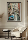 Een ingelijst schilderij Kanaal in Venetië van CollageDepot is op een beige muur gemonteerd. Daaronder staan een moderne witte fauteuil en een rond zwart bijzettafeltje met een boek erop. Licht stroomt de kamer binnen en creëert een rustige en uitnodigende sfeer. Deze elegante wanddecoratie voegt verfijning toe aan de ruimte.