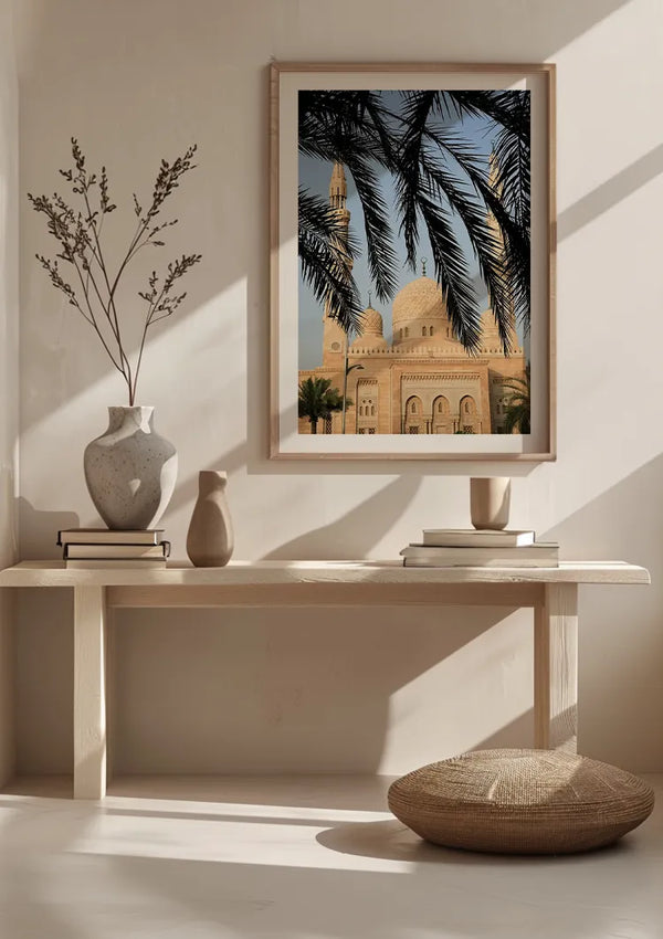 Een minimalistische interieurscène met een lichte houten bank met keramische vazen, een kussen op de vloer en een ingelijst Moskee Met Sierlijke Structuur Schilderij van CollageDepot van een architectonisch bouwwerk met koepels en minaretten gezien door palmbladeren, hangend aan de muur boven de bank met een magnetisch ophangsysteem.,Lichtbruin