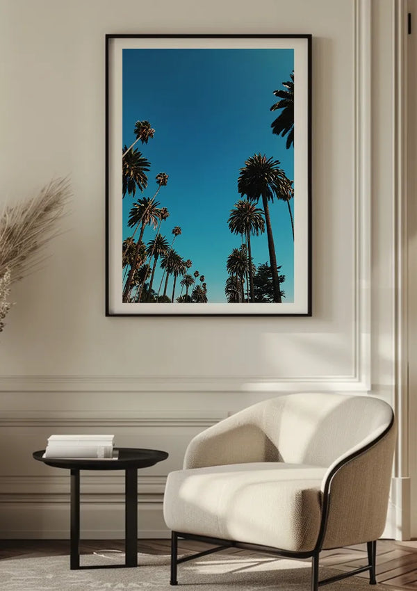 Het gezellige interieur is voorzien van een ingelijste foto van hoge palmbomen tegen een helderblauwe lucht. Onder het kunstwerk creëren een stoel met witte kussens en een klein rond zwart tafeltje met een stapel boeken een serene en uitnodigende leeshoek, waaronder de stijlvolle bbb 023 - natuur van CollageDepot.,Zwart