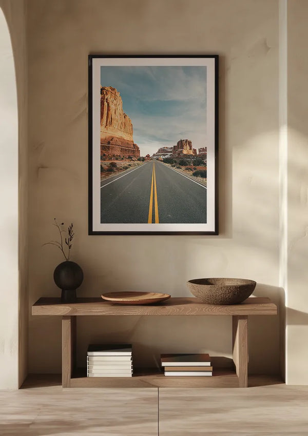 Een ingelijst Canyon road schilderij van CollageDepot hangt aan een beige muur als elegante wanddecoratie. Hieronder staat een houten consoletafel met een minimaal decor, waaronder een zwarte vaas met een enkele tak, een bruine kom en een paar gestapelde boeken.