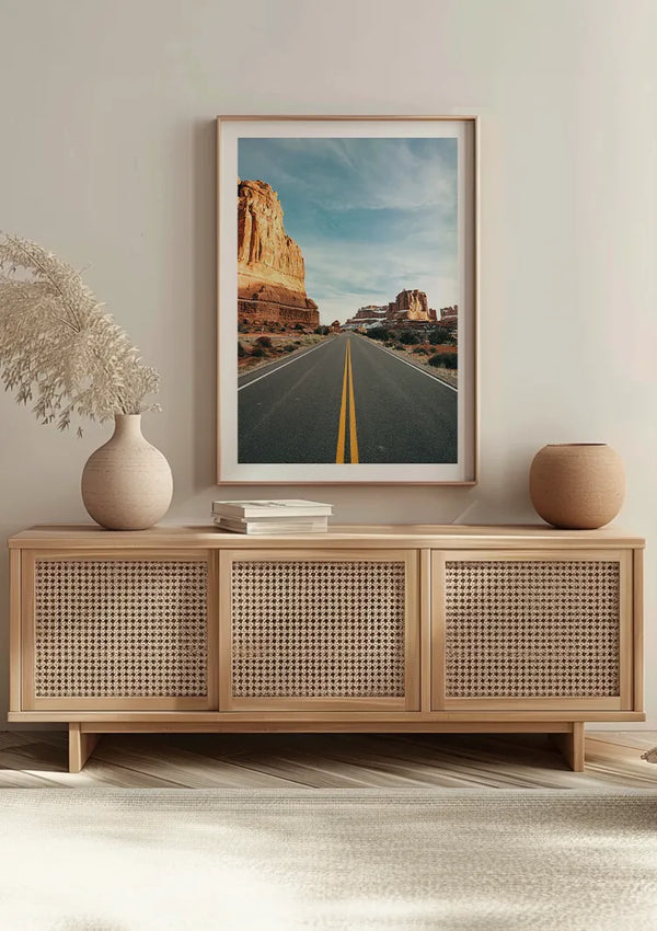 Het minimalistische interieur omvat een lichtgekleurde houten console met drie kastdeuren met een geweven rotanontwerp. Daarop staan vazen en boeken. Hierboven is een grote ingelijste foto van een woestijnweg met rotsformaties opgehangen met een Canyon road-schilderij van CollageDepot voor naadloze wanddecoratie.