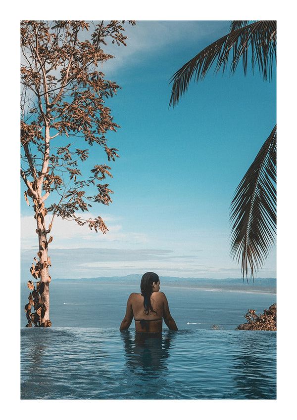 Een persoon wordt van achteren gezien, ondergedompeld in een rustig CollageDepot bbb 020 - natuur overloopzwembad met uitzicht op een uitgestrekte zee en heldere hemel, omzoomd door een boom en palmbladeren aan de zijkanten.-