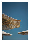 Close-up van een geweven parasol tegen een helderblauwe lucht, die doet denken aan een zomers schilderij. Alleen het bovenste deel van de paraplu is zichtbaar, met gedeeltelijke weergaven van andere parasols op de achtergrond. De afbeelding heeft een verticale oriëntatie, perfect voor wanddecoratie met een magnetisch ophangsysteem. Maak kennis met het schilderij De Geweven Parasols van CollageDepot.-