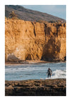 Een persoon in een wetsuit staat op een surfplank en rijdt op een kleine golf nabij een rotsachtige kustlijn met ruige kliffen op de achtergrond. Bovenop de kliffen zijn enkele palmbomen en struiken zichtbaar, die lijken op een pittoresk schilderij Surfen bij kliff van CollageDepot. De lucht is helder en blauw.