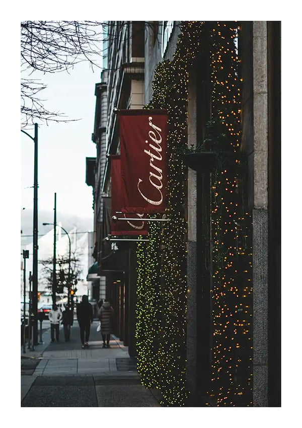 Een straatbeeld in de stad toont een Cartier-winkel met een verlichte gevel bedekt met groen licht. Boven de ingang hangt een rood bord met het Cartier-logo. Bomen met kale takken omzomen het trottoir en in de verte lopen een paar voetgangers, waarvan er één een nabijgelegen CollageDepot Cartier-winkel bewondert die met behulp van een magnetisch ophangsysteem schildert.