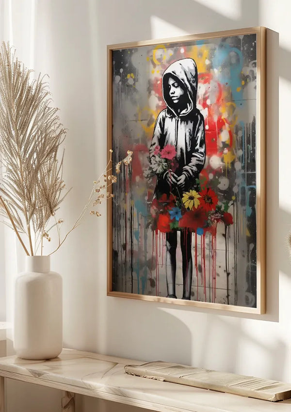 Een ingelijst schilderij, dat doet denken aan graffitikunstwerk, toont een persoon die een kledingstuk met capuchon draagt en kleurrijke bloemen vasthoudt. De achtergrond is een mix van grijstinten met spatten van levendige kleuren. Het CollageDepot Graffiti Met Jongen En Bloemen Schilderij wordt getoond op een witte muur boven een wit vlak met een plant in een vaas.,Lichtbruin