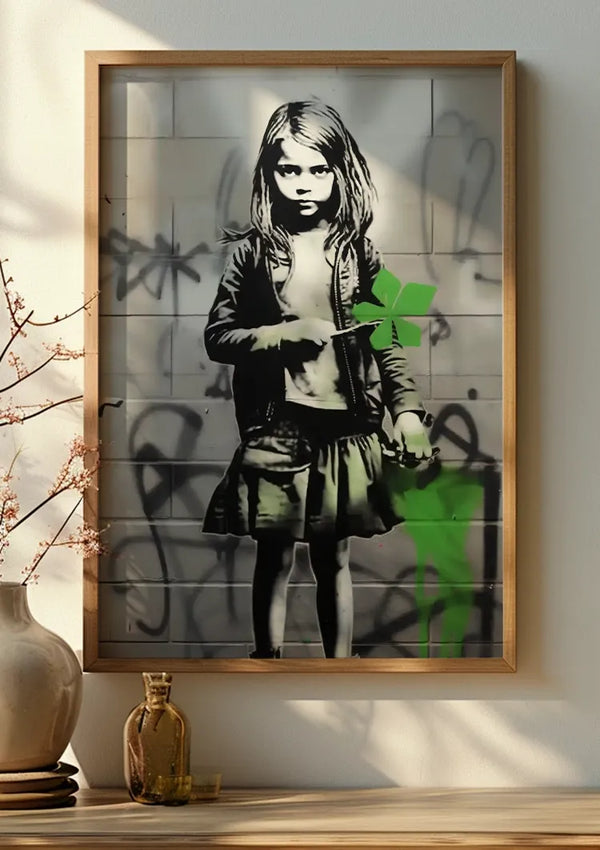 Een ingelijst CollageDepot Graffiti Girl Klavertje Vier Schilderij toont een zwart-wit stencil van een jong meisje dat een groene origamivogel vasthoudt. Ze staat tegen een met graffiti bedekte muur, gekleed in een jasje en een rok. Deze wanddecoratie wordt aan de muur gehangen met een vaas en pot op een tafeltje ernaast, met behulp van een onzichtbaar magnetisch ophangsysteem.