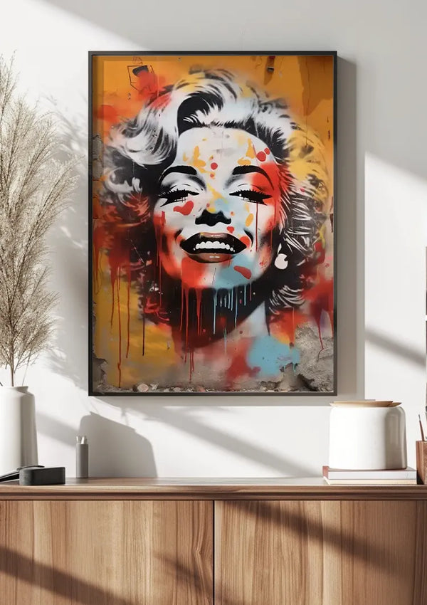 Een ingelijst Graffitikunstwerk Van Marilyn Monroe Schilderij van CollageDepot met levendige verfspatten wordt aan een witte muur boven een houten kast gehangen. De kast bevat minimalistische decorartikelen, waaronder een potplant, een boek en een cilindrische container.,Zwart