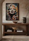 Een ingelijste, kleurrijke Marilyn Monroe Met Levendige Kleuraccenten Schilderij van CollageDepot hangt aan een beige muur boven een houten consoletafel. Op de tafel staan diverse decoratieve voorwerpen waaronder schalen, een vaas met takken en een stapel boeken. De kamer is voorzien van neutraal gekleurde vloeren en verfijnde wanddecoratie.,Zwart