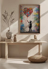 Een ingelijst kunstwerk, het Kind Reikt Naar Hartvormige Ballon Schilderij van CollageDepot, wordt tentoongesteld op een lichthouten tafel. De tafel is versierd met twee vazen en verschillende gestapelde boeken. Vooraan ligt een rond kussen op de grond.,Lichtbruin