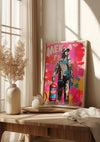 Een ingelijst portret in stedelijke stijl met een kleurrijke graffiti-achtergrond rust op een houten tafel met een neutraal getint decor. Het kunstwerk toont een staande figuur met verfspatten en het woord "MEK" zichtbaar in grote letters. Zonlicht filtert door hoge ramen naar binnen, waardoor opvallende Vrolijke Graffitistijl Met Man Schilderij van CollageDepot ontstaat.,Lichtbruin