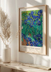 Een ingelijste poster van CollageDepot’s V. Van Gogh Paarse Irissen Schilderij wordt tentoongesteld als een prachtig stukje wanddecoratie. Levendige blauwe irissen met groene bladeren boeien het oog. Geplaatst boven een witte plank met een vaas met gedroogde takken, wordt het tafereel prachtig verlicht door zonlicht.,Lichtbruin