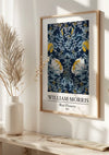 Een ingelijst kunstwerk met de titel "W. Morris Blue Flowers Schilderij" van CollageDepot uit 1874 wordt elegant weergegeven als een prachtige wanddecoratie aan de muur. De botanische illustratie toont blauwe en gele bloemen, met droog gras in een keramische vaas op een witte plank eronder, wat de natuurlijke sfeer versterkt.,Lichtbruin
