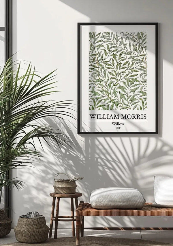 Een ingelijst "W. Morris Willow Schilderij" van CollageDepot hangt aan een zonverlichte muur, dankzij een magnetisch ophangsysteem. Onder het kunstwerk staat een rieten bankje met kussens en een witte deken. Op en naast de bank staan twee rieten manden, terwijl links een grote potplant staat.,Zwart