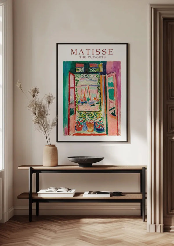 Een ingelijst kleurrijk schilderij getiteld "Matisse - The Cut Outs Open Window Schilderij" van CollageDepot hangt aan een lichtgekleurde muur met behulp van een magnetisch ophangsysteem boven een minimalistische houten consoletafel. Op de tafel staan een kom, een vaas met gedroogde bloemen en een open boek, allemaal in een kamer met een houten vloer met visgraatmotief.,Zwart