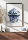 Aan een witte muur hangt een wanddecoratie met een ingelijst Delfts Blauw Cupcake Schilderij van CollageDepot. Hieronder staat een houten plank met een theepot met witte en blauwe bloemen en gedroogde planten in een witte vaas. Natuurlijk licht werpt schaduwen op de muur.,Zwart