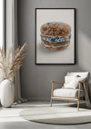 Een moderne woonkamer is voorzien van een groot ingelijst Delfts Blauw Keramische Hamburger Schilderij van CollageDepot met een afbeelding van een hamburger met sesamzaadjes en blauw-witte porseleinen elementen. De ruimte omvat een stoel met witte kussens, een beige rond tapijt en een hoge vaas met gedroogd pampagras, allemaal aangevuld met strakke wanddecoratie-opties voor extra stijl.,Zwart