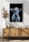 Aan een witte muur boven een houten dressoir hangt een ingelijst kunstwerk van een koe met een blauw-wit bloemmotief. Het dressoir is versierd met twee vazen en enkele gedroogde planten. Zonlicht werpt schaduwen op de muur en de vloer.,Zwart