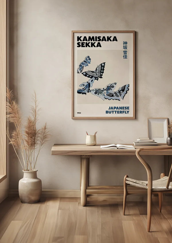 Tegen een muur staat een houten bureau met een stoel. Boven het bureau hangt een ingelijste poster met de tekst "K. Sekka Japanese Butterfly Schilderij" en "Japanese Butterfly" samen met illustraties van vlinders, wat een artistiek tintje geeft aan de wanddecoratie. Naast het bureau staat een vaas met gedroogde planten.,Lichtbruin
