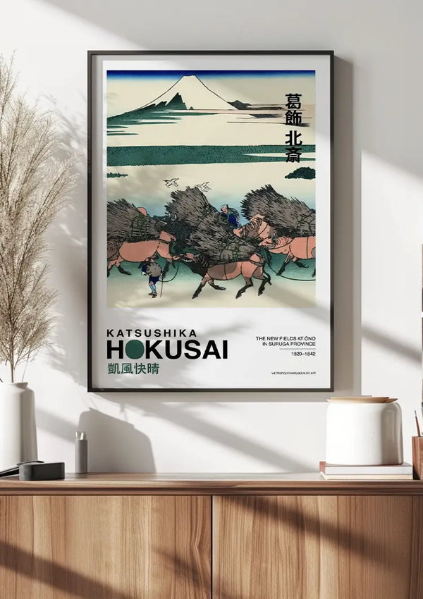 Een ingelijst schilderij van K. Hokusai The New Fields at Ono in de provincie Suruga van CollageDepot hangt aan een muur boven een houten kast. Het beeld, dat dient als schitterende wanddecoratie, toont verschillende paarden en mensen die goederen dragen met de berg Fuji op de achtergrond. De titel en de naam van de kunstenaar worden prominent weergegeven op de onderkant van de poster.,Zwart