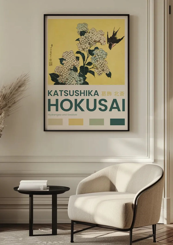Een ingelijste poster met de titel "K. Hokusai Hydrangea And Swallow Schilderij" van CollageDepot met een illustratie van hortensia's en een zwaluw hangt aan de muur boven een beige fauteuil en een kleine ronde zwarte tafel. De poster wordt veilig weergegeven met een magnetisch ophangsysteem en bevat ook drie verfstalen en tekst in zowel het Engels als het Japans.,Zwart