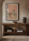 Een ingelijste print met de titel "S. Harunobu The Water Vendor Schilderij", een schilderij van CollageDepot, hangt aan een beige muur boven een houten consoletafel. De tafel is voorzien van twee houten schalen en een keramische vaas met textuur. Aan het rechteruiteinde van de consoletafel wordt een tak in een vaas geplaatst.,Zwart