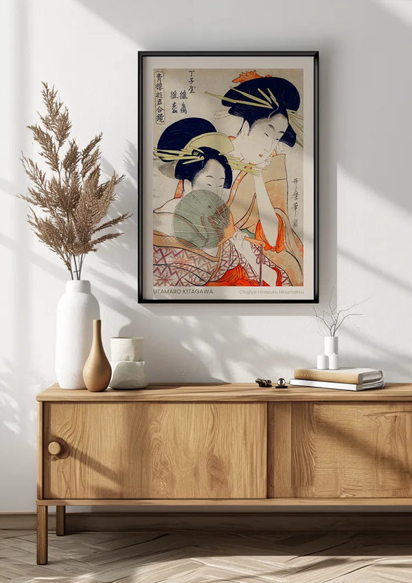 Een ingelijste U. Kitagawa Chojiya Hinazuru Hinamatsu Schilderij van CollageDepot hangt aan een witte muur boven een houten dressoir. Het dressoir heeft een beige vaas met gedroogde planten en een kleinere witte vaas. Deze wanddecoratie draagt bij aan de minimalistische esthetiek van de kamer, met natuurlijk licht dat schaduwen werpt.,Zwart