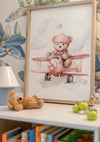 Op een plank staat een ingelijst CollageDepot Teddybeer In Roze Vliegtuig Schilderij van een teddybeer met een pilotenhoed. De teddybeer zit op een rood speelgoedvliegtuig. Op de plank staan ook een kleine teddybeer, een witte lamp, speelgoed en kinderboeken. De achtergrond heeft bloemenbehang.,Lichtbruin
