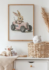 Een kinderkamer is voorzien van een ingelijste afbeelding van een konijn dat in een roze speelgoedauto rijdt met 'kinder' op de zijkant geschreven, moeiteloos opgehouden door een elegant magnetisch ophangsysteem. Onder de foto staat een ladekast met een rieten mand met daarin een pluchen kat en diverse andere knuffels. Gedroogd gras siert de hoek.Productnaam: Konijn in Roze Auto SchilderijMerknaam: CollageDepot,Lichtbruin