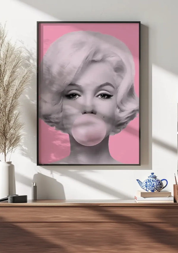 Een ingelijst zwart-wit portret van een vrouw met kort golvend haar die een kauwgombel blaast, hangt als opvallende wanddecoratie aan een witte muur. De achtergrond van het Bubblegum Beauty Popart Schilderij van CollageDepot is roze. Op de voorgrond zijn een plant, theepot en houten kast zichtbaar.