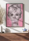 Een ingelijst Bubble Gum Bliss Popart Schilderij van CollageDepot van een vrouw die kauwgom blaast, wordt aan een witte muur gehangen met behulp van een magnetisch ophangsysteem. De achtergrond is roze. Onder het frame, dat dient als opvallende wanddecoratie, bevindt zich een houten oppervlak versierd met decoratieve items zoals een blauwe theepot, beker en vaas met gedroogd pampagras.