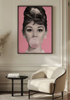 Een wanddecoratie met het Pink Bubble Gum Lady Popart Schilderij van CollageDepot is gemonteerd op een beige muur. Onder het schilderij staat een moderne witte fauteuil en een klein zwart bijzettafeltje met een boek. Een gedroogde plant siert de hoek van de kamer.