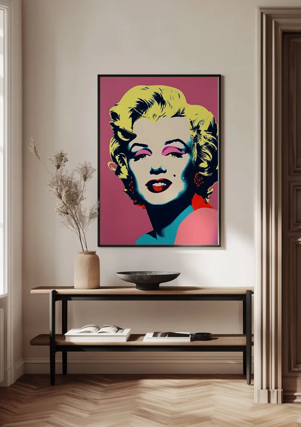 Een ingelijst iconisch Marilyn Monroe Popart Schilderij van CollageDepot met blond haar hangt aan een lichtgekleurde muur boven een houten consoletafel. Op de tafel staat een keramieken vaas met gedroogde takken, een donkere kom en een open boek. Deze wanddecoratie versterkt de minimalistische esthetiek van de setting.