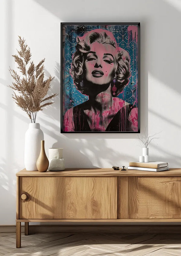 Aan een witte muur boven een licht houten dressoir hangt een Marilyn Monroe Spetterende Expressie Popart schilderij van een vrouw met kort blond haar en felrode lippen. Op het dressoir staat een hoge witte vaas met gedroogde planten, een witte mok en diverse andere decoratieve items, allemaal beveiligd door een innovatief magnetisch ophangsysteem van CollageDepot.